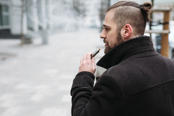 Men with beard hold and smoke his electronic cigarette outdoor in black, grey backgraund