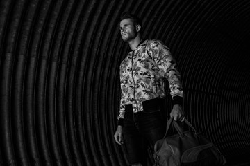 Fototapeta na wymiar Sexy muscular young model with beard on dark background. Fashion portrait of brutal strong guy in trendy clothes and hairstyle. Fashion concept. Black and white. Stylish man traveling with a bag.