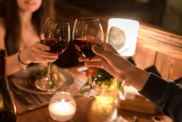Hand of man and woman sitting in a dark restaurant by a table and holding glasses with red wine