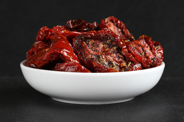 Sun-dried tomatoes in sunflower oil with spices (rosemary, garlic, basil, black pepper) in a white ceramic bowl, standing on a black stone surface against a black stone wall