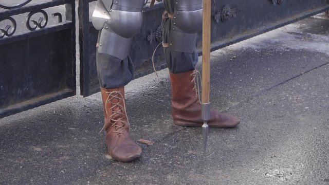 A knight in a metal suit and brown leather boots stands near an iron gate holding a spear in his hand