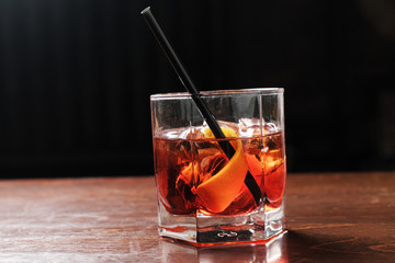Glass of a Boulevardier cocktail with big ice cube and orange zest on the steel wooden bar counter on the blurred background