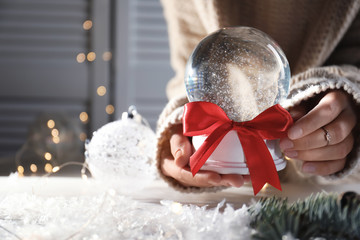 Woman holding Christmas snow globe with red bow on blurred background, closeup. Space for text