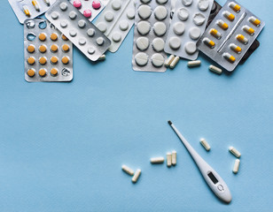 White, yellow, pink tablets in blisters and thermometer on a blue background top view. Medical pharmacy concept. Traditional medicine. Copy space.