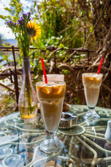 Two Ice coffee glasses with red drinking straw, vase with flowers, ashtray and red candle on the metal glass table surrounded by exotic plants. Romantic place.