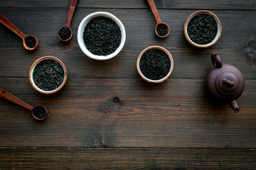 Set of dry tea leaves in bowls and spoons near tea pot for tea ceremony on dark wooden background top view copy space