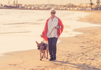 Happy attractive senior woman with her german shepard dog walking on the beach at autumn sunset