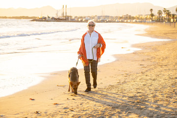 Happy attractive senior woman with her german shepard dog walking on the beach at autumn sunset