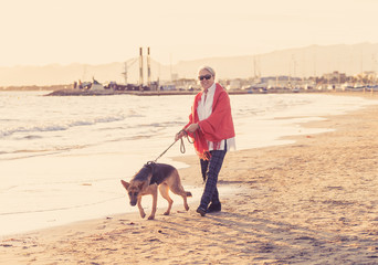 Happy attractive senior woman with her german shepard dog walking on the beach at autumn sunset