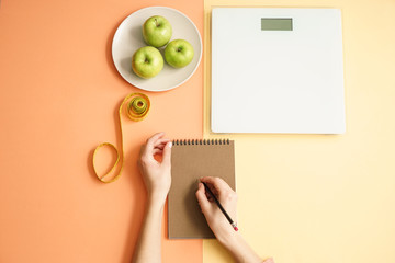 Obraz premium Woman writing in notebook, scales, apples and measuring tape on color background. Weight loss concept