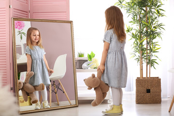 Cute little girl with teddy bear looking in mirror at home