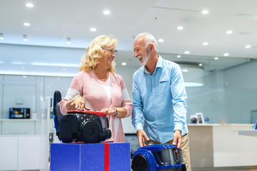 Smiling married old couple can't decide which vacuum cleaner to buy. Tech store interior.