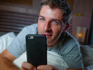 lifestyle home portrait of young attractive and relaxed man using internet social media app on mobile phone in his bedroom late at night lying on bed networking happy