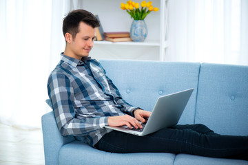 A Young attractive guy man is browsing at his laptop, sitting at home on the cozy blue sofa at home, wearing casual outfit
