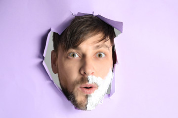 Surprised man with shaving foam on his face visible through hole in torn color paper