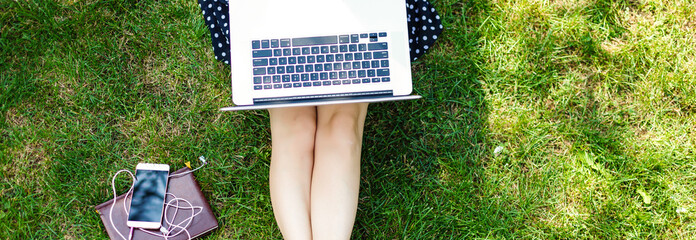 Top view of woman sitting in park on the green grass with laptop, notebook and smartphone, hands on keyboard. Computer screen mockup. Student studying outdoors. Copy space for text