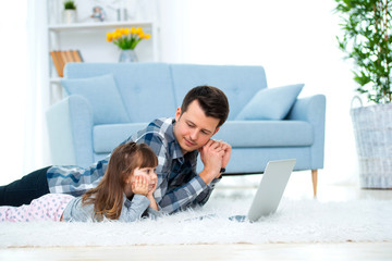 Cute little girl, daughter, sister and young dad father or brother look at the laptop monitor computer, lying on carpet on the floor in living room