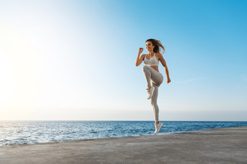 Fototapeta na wymiar Young motivated happy sporty female runner fitness instructor, jumping, excercise near sea enjoying fresh morning air training, smiling facing sunlight, workout wearing activewear, running in quay