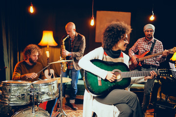 Mixed race woman singing and playing guitar while sitting on chair with legs crossed. In background drummer, saxophonist and bass guitarist.