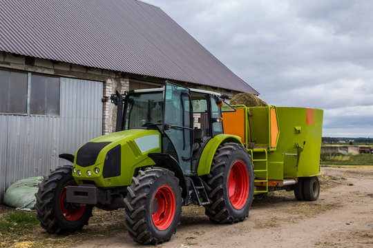 Tractor with a trailer for mixing and distribution of feed for cows. Behind the tractor is a barn and fields. Necessary equipment for a dairy farm.