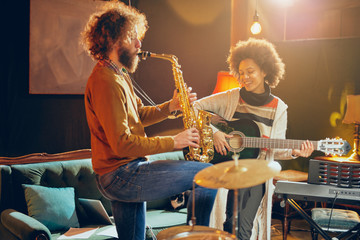 Mixed race woman playing acoustic guitar while man playing saxophone. Home studio interior.