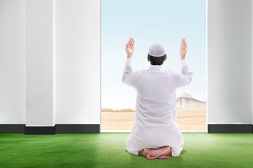 Back view of asian muslim man sitting in praying position on the carpet raise the hand and gazing...