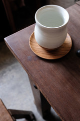 White ceramic asian drinking cup on wooden coaster on rustic dark weathered wooden table top on blurred background with empty copy space. Minimal home interior decoration concept.