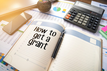 Business accessories, smartphone calculator, laptop, reports and diary with text How to get a grant