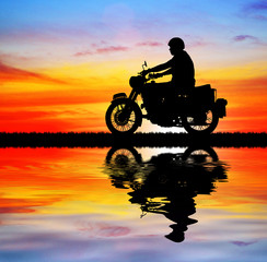 Fototapeta premium Silhouette biker with his motorbike beside the natural lake and beautiful sunset sky.