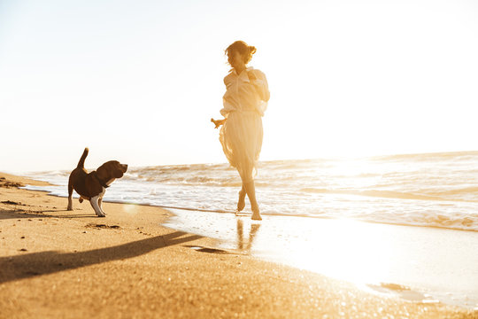 Image of positive woman 20s playing with her dog, while walking by seaside