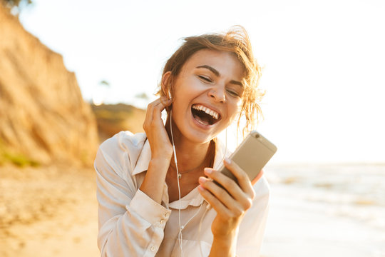 Image of excited girl 20s wearing earphones laughing and using mobile phone, while walking by seaside
