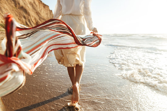 Portrait of positive girl 20s walking with waving scarf, along seashore