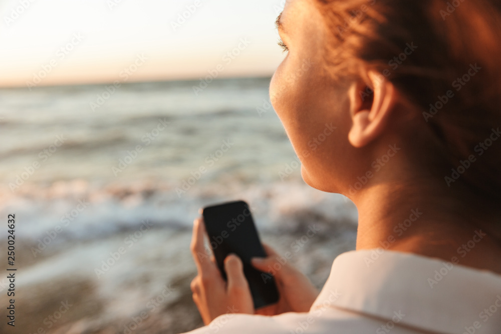 Poster Image of young woman 20s using smartphone and looking at water, while walking by seaside