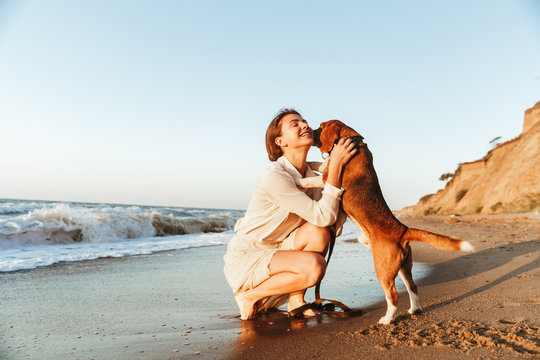 Image of happy woman 20s hugging her dog, while walking along the beach