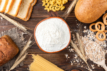 Wheat products on dark wooden background. Bread, spaghetti, wheat flour and flakes, loaf and dryings. Carbohydrates.