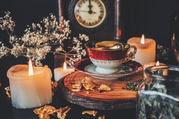 Making a cup of tea in witch's kitchen. A vintage red white and gold colored teacup placed on a wooden platform on a black table. Kitchen witchcraft with tea, herbs and spices with burning candles