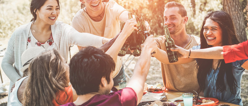 Happy millennial friends cheering with beers at barbecue dinner party outdoor - Focus on guys faces