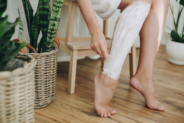 Hair Removal concept, depilation process. Young woman in white towel applying shaving cream on her legs and holding holding plastic razor in home bathroom with green plants. Skin care - Powered by Adobe