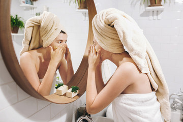 Young happy woman in towel applying organic face scrub and looking at round mirror in stylish...