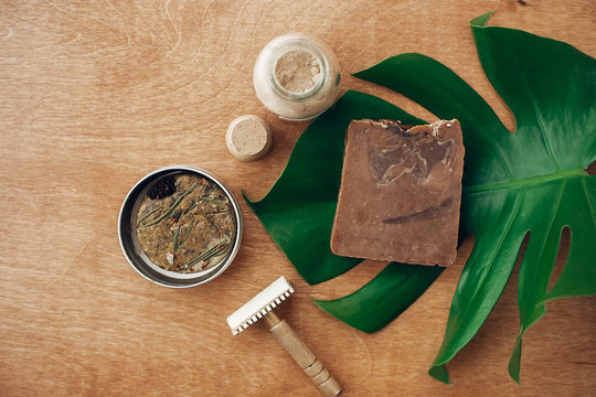 Zero waste concept, flat lay. Natural soap, solid shampoo in metal tin, reusable razor, ubtan on wooden background with green monstera leaf. Plastic free beauty essentials. Ban plastic