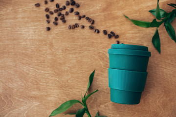 Stylish reusable eco coffee cup  on wooden background with coffee beans and green bamboo leaves....