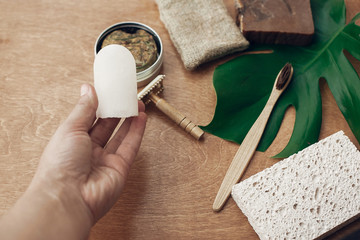 Hand holding natural eco crystal deodorant on background of bamboo toothbrush, soap, solid shampoo, metal razor, sponge on wood with green monstera leaf.  Zero waste concept. Sustainable lifestyle