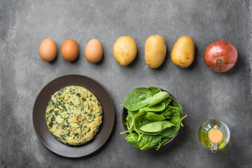 Homemade delicious frittata with spinach on plate. Recipe ingredients potatoes eggs olive oil in bottle on dark concrete background. Knolling lay out. Rustic style. Overhead top view copy space