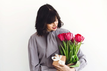 Beautiful brunette girl holding stylish bouquet of tulips and gift box on white background indoors, space for text. Happy young woman with flowers. Happy mothers day. International womens day