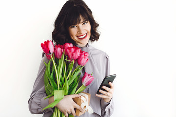 Beautiful happy girl with bouquet of tulips and gift box on white background indoors, space for text. Stylish young woman holding phone and flowers.  International womens day. Hello spring.