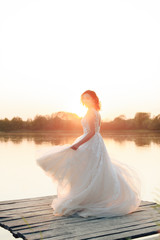 Fototapeta na wymiar young beautiful bride in white dress on wooden pier near water at sunset