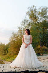 Fototapeta premium young beautiful bride in white dress on wooden pier near water at sunset