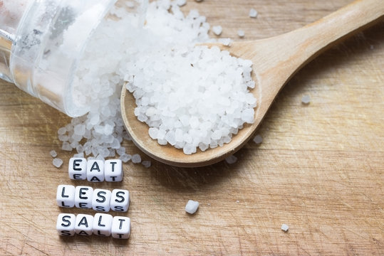 Eat less salt advice written with plastic letter beads on wooden cutting board, near granulated salt &ndash; healthy food lifestyle 