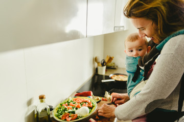 Busy mother preparing food in the kitchen while taking care of her baby, in a baby carrier using the kangaroo method.