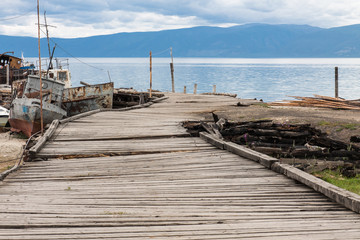 Fototapeta premium The cemetery of boats. Abandoned rusty boats on the shore near old broken wooden pier. In the background blue sky with clouds, lake and mountains